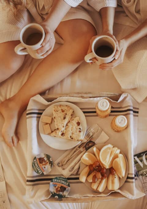 Two people sit on a bed wearing robes, holding coffee mugs. Between them is a breakfast tray with pastries, fruit, cheese, and yogurt. A map and a magazine are also on the bed.