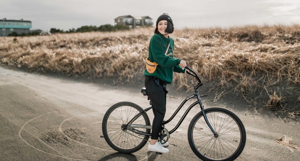 A person wearing a green sweater, black pants, and a helmet is smiling while standing with a black bicycle on a paved path. In the background, there are brown fields and a cloudy sky.