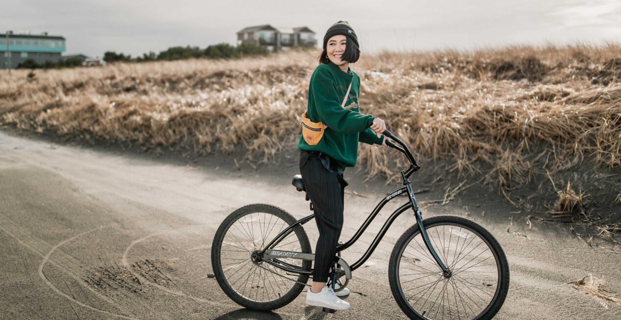 A person wearing a green sweater, black pants, and a helmet is smiling while standing with a black bicycle on a paved path. In the background, there are brown fields and a cloudy sky.