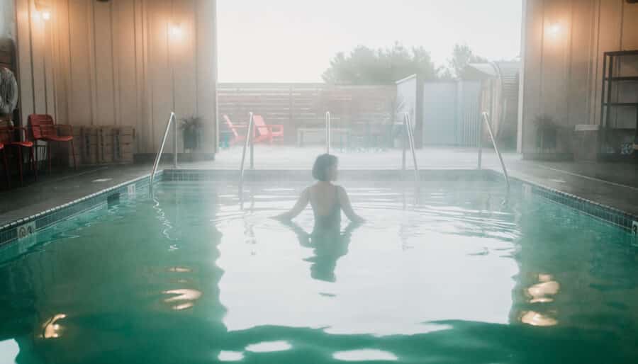 A person stands waist-deep in an indoor swimming pool, facing a large open entrance that leads to an outdoor area with patio chairs. The atmosphere is misty, and soft lighting illuminates the space.