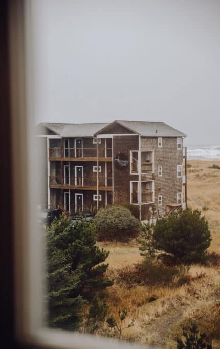 View of a three-story, weathered wooden building with "Discovery Coasters" signage, surrounded by grass and trees. The scene is overcast, with the ocean visible in the background. The image is taken from a window perspective.