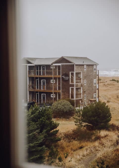 View of a three-story, weathered wooden building with "Discovery Coasters" signage, surrounded by grass and trees. The scene is overcast, with the ocean visible in the background. The image is taken from a window perspective.