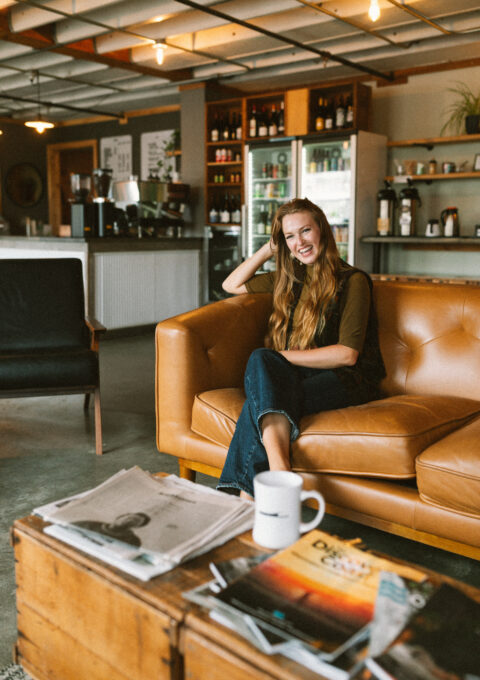 A woman with long hair sits on a brown leather couch in a cafe, smiling at the camera. A wooden coffee table with newspapers, a magazine, and a white mug is in the foreground. Shelves and a fridge are in the background.