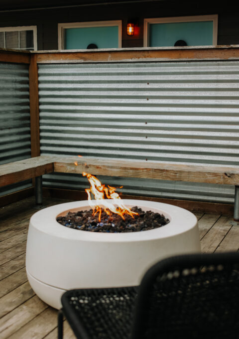 A circular fire pit is on a wooden deck, surrounded by a metal bench and corrugated metal privacy screen. A small, contained flame burns in the center of the pit. A red light is mounted on the wall behind.