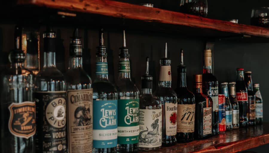 A wooden bar shelf displays various liquor bottles, including vodka, gin, rum, whiskey, liqueurs, and mixers. Some glasses and barware are also visible on the upper shelf against a dark background.