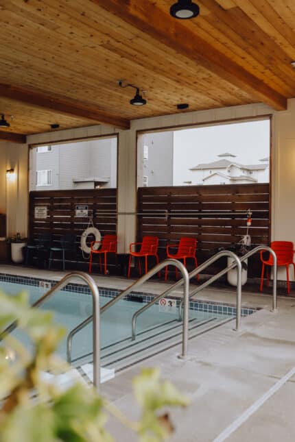 Indoor swimming pool area with wooden ceiling and large windows. The pool is bordered by silver handrails and surrounded by red chairs against a wooden fence. Life preservers are mounted nearby. Outside, buildings are visible through the windows.