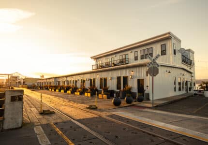 A large white building with balconies and potted plants sits beside railroad tracks at sunset. A railroad crossing sign is visible in front, with warm sunlight illuminating the scene.