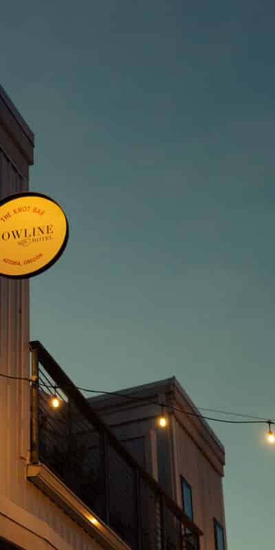 Exterior of a building at dusk with a round lit sign reading “BOWLINE EAST BAY” and string lights hanging across the sky. The building has metal siding and a visible balcony.
