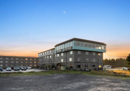 A multi-story hotel building is shown at sunset with a crescent moon in the sky. Several cars are parked in the front lot. The building has large windows and a mix of neutral tones.