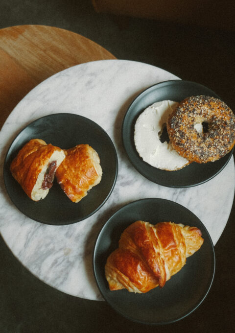 A small round marble table with three black plates. One plate has a croissant, another has a pastry filled with red jam, and the third has a bagel topped with seeds and cream cheese. A wooden chair is partially visible next to the table.