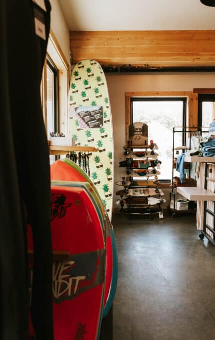 A surf shop interior showcasing a selection of surfboards, skateboards, and clothing. A prominent surfboard with a pineapple pattern leans against a wall. Sunlight filters through the windows, illuminating the boards and clothes on display.