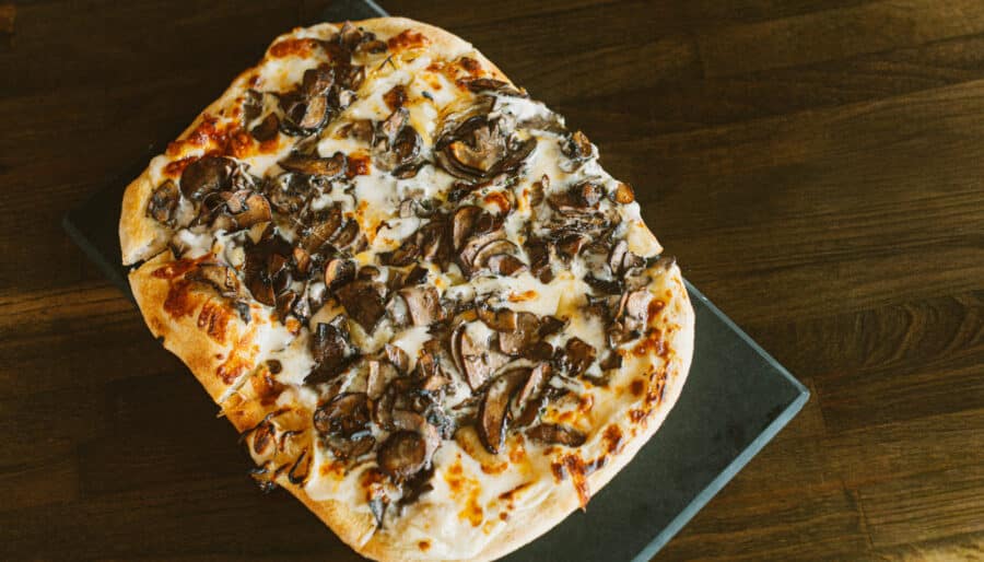 A rectangular flatbread pizza topped with mushrooms and melted cheese sits on a dark rectangular serving board on a wooden table.