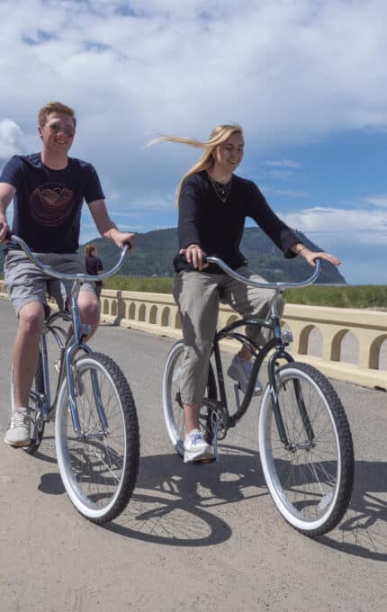 A man and a woman are riding bicycles on a paved path along a beach. The sky is partly cloudy, and the ocean is visible in the background. Both are wearing casual clothing, and a concrete railing lines the path.