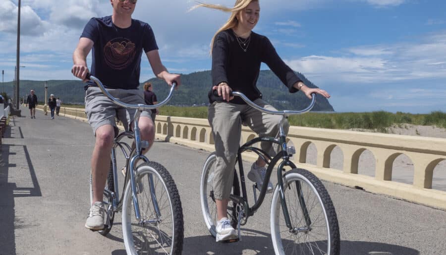 A man and a woman are riding bicycles on a paved path along a beach. The sky is partly cloudy, and the ocean is visible in the background. Both are wearing casual clothing, and a concrete railing lines the path.