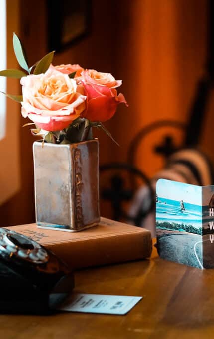 A vintage rotary phone is placed on a wooden table next to a vase with pink roses. A book is stacked beneath the vase. Beside them, a greeting card reads "Hi there, we're glad you're here." The scene is warmly lit by natural light.