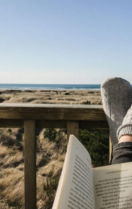 A person wearing gray socks is relaxing on a wooden deck, holding an open book. The view overlooks a grassy dune landscape with the ocean and a clear blue sky in the background.