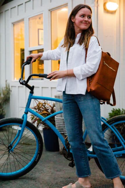 A woman stands next to a blue bicycle outside a building. She is wearing a white shirt, blue jeans, and sandals, with a brown backpack over her shoulder. The background features a lit window and potted plants.