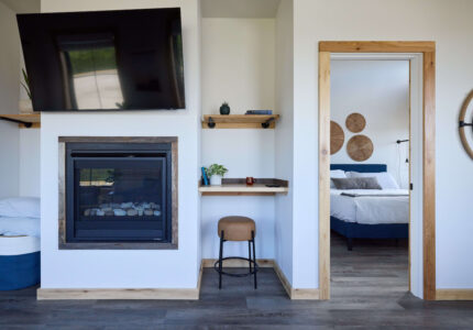 A modern room with a wall-mounted TV above a fireplace, a small desk with a stool, and a door opening to a bedroom with a bed and decorative wall pieces. The room has wood trim and gray flooring.