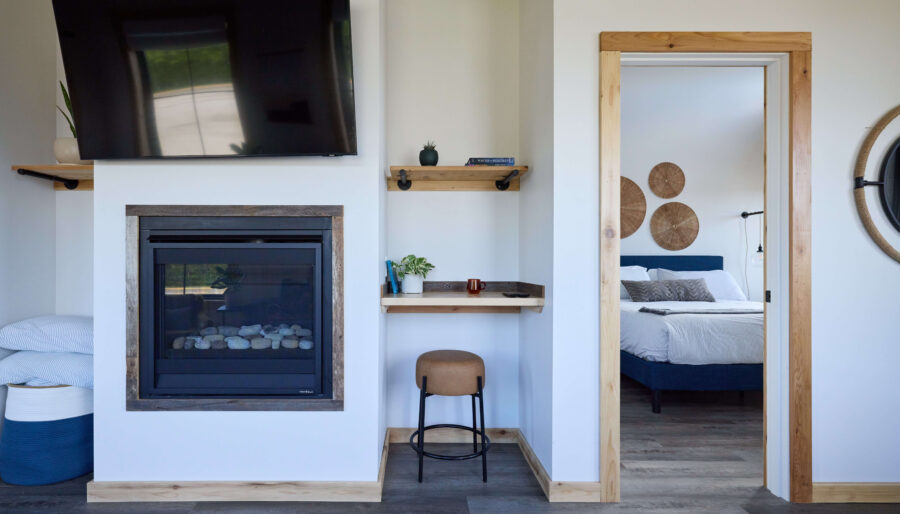 A modern room with a wall-mounted TV above a fireplace, a small desk with a stool, and a door opening to a bedroom with a bed and decorative wall pieces. The room has wood trim and gray flooring.