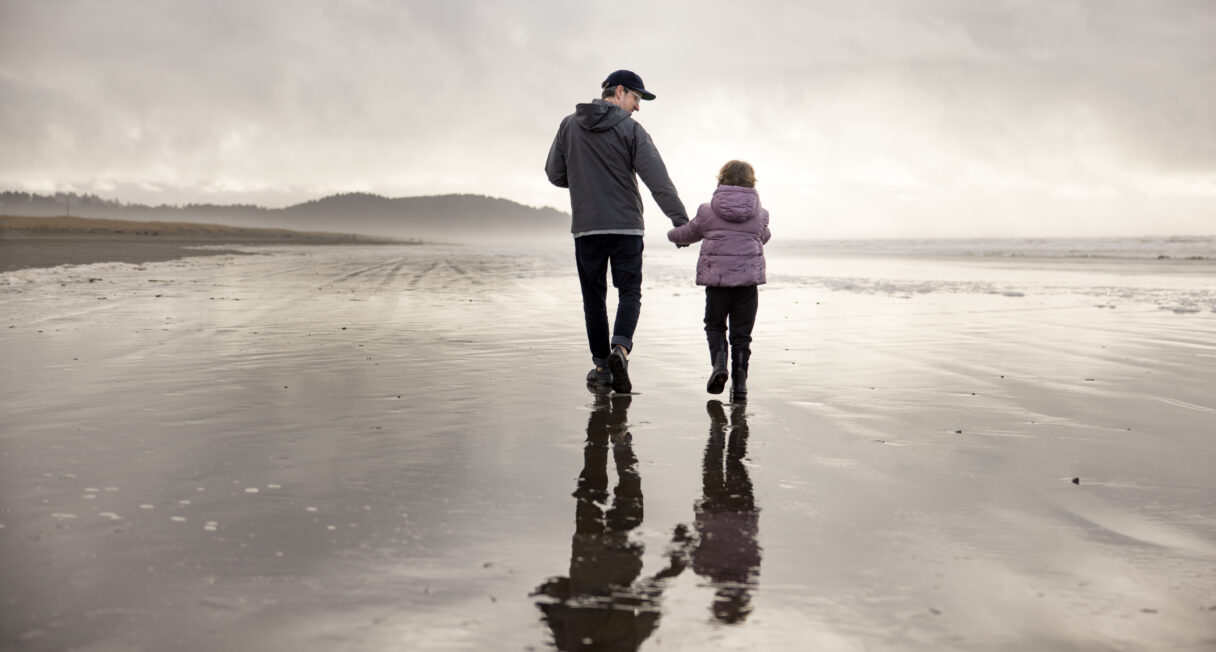 A person and a child walk hand in hand along a reflective wet beach, both dressed in jackets. The sky is overcast, and the horizon shows a distant line of trees. The beach is wide and empty, creating a serene atmosphere.