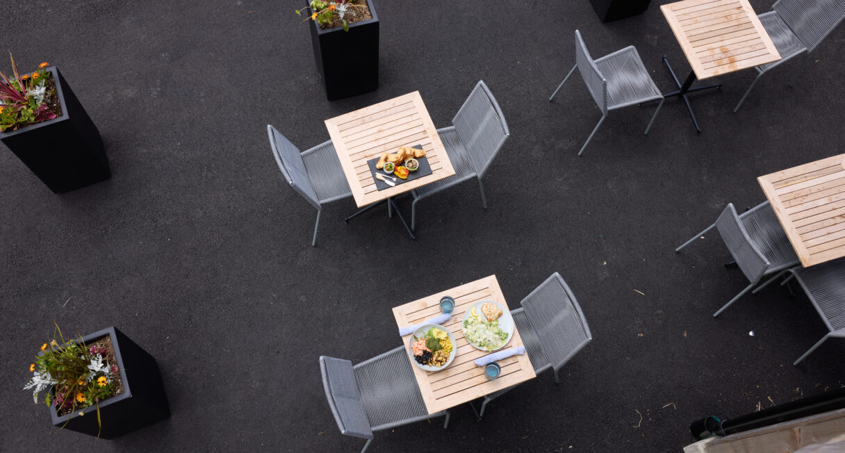 Outdoor seating area with wooden tables and metal chairs on a dark pavement. Two tables feature plates of food and drinks. Potted plants are placed nearby, adding greenery to the setting. Overhead view capturing a casual dining environment.