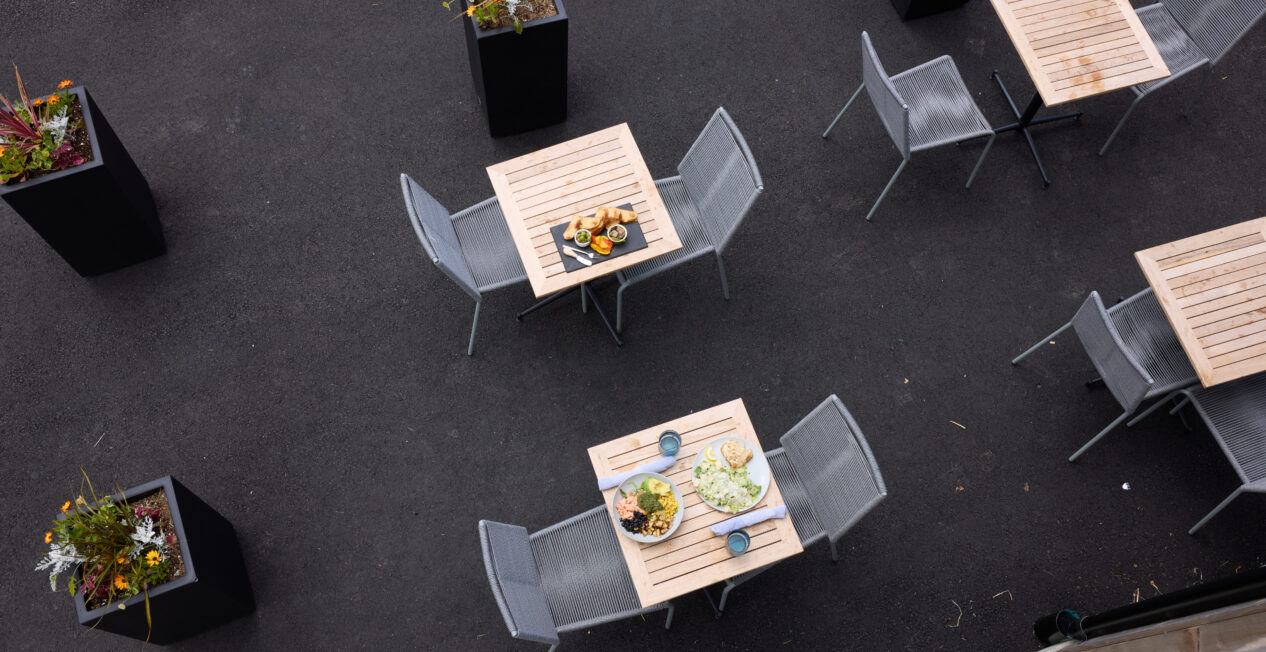 Outdoor seating area with wooden tables and metal chairs on a dark pavement. Two tables feature plates of food and drinks. Potted plants are placed nearby, adding greenery to the setting. Overhead view capturing a casual dining environment.