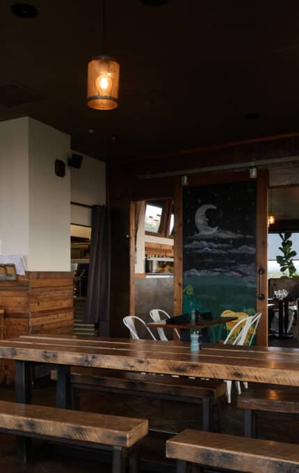 A rustic restaurant interior with wooden benches and tables. The bar to the left has shelves of bottles. Hanging lights create a warm ambiance. Artwork of a night sky decorates a wall. A few patrons are seated at tables in the distance.