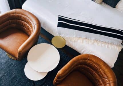 A top-down view of a chic interior with two brown leather armchairs, a small white round coffee table, and part of a bed with a black and white striped throw blanket.