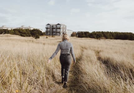 A person wearing a striped shirt and jeans walks through a grassy field toward a distant row of buildings under a partly cloudy sky.