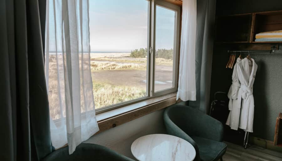A hotel room with two dark green chairs and a small round white table by a large window overlooking a grassy landscape; an open closet with robes and towels is visible on the right.
