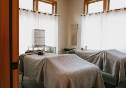Two massage tables covered with textured gray blankets are set up side by side in a bright room with white curtains, wooden window frames, and a small cabinet. A mug and towels are placed on each table.