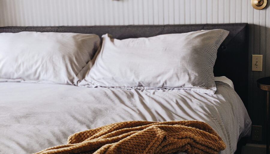 A neatly made bed with striped bedding and two pillows sits against a gray paneled wall. A yellow textured blanket is draped at the foot of the bed. A diamond-shaped mirror and a wall lamp are visible above the bed.
