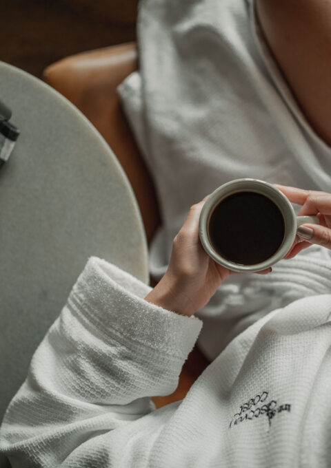 A person in a white bathrobe sits in a chair holding a cup of coffee. A coffee pot and a small container are placed on a round table with a checkered cloth beside it. The scene suggests a moment of relaxation or a morning routine.