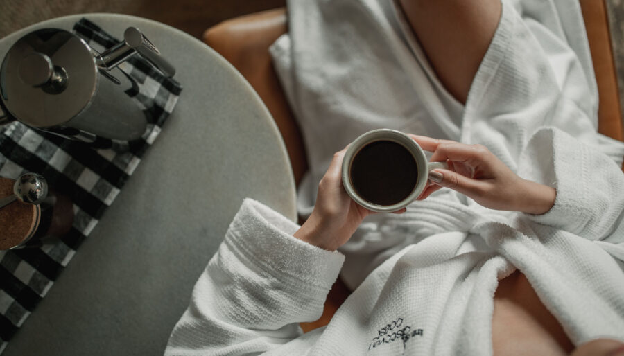 A person in a white bathrobe sits in a chair holding a cup of coffee. A coffee pot and a small container are placed on a round table with a checkered cloth beside it. The scene suggests a moment of relaxation or a morning routine.
