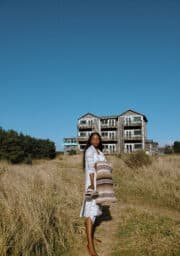 A woman stands on a grassy path leading to a two-story house. She is holding a patterned blanket and wears a light-colored dress. The sky is clear and blue, and there are trees on the left side of the scene.