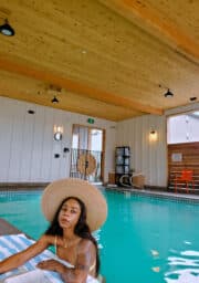 A person wearing a large straw hat sits by the edge of an indoor pool with a wooden ceiling. They are holding a book and sitting on a striped towel. A door and several chairs are visible in the background.