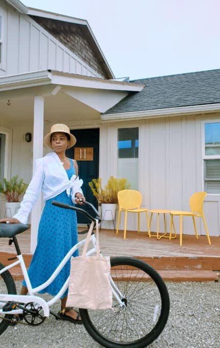 A person stands in front of a house holding the handlebars of a white bicycle. They are wearing a blue dress, white shirt, and straw hat. The house has a porch with a yellow table and chairs, and potted plants are placed nearby.