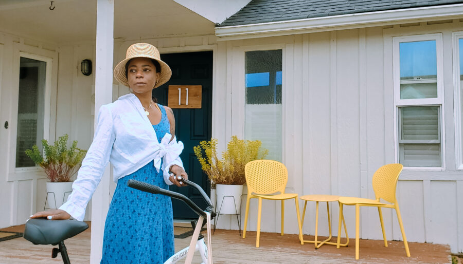 A person stands in front of a house holding the handlebars of a white bicycle. They are wearing a blue dress, white shirt, and straw hat. The house has a porch with a yellow table and chairs, and potted plants are placed nearby.