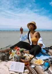 A person sits on a picnic blanket at a beach. They wear a hat, yellow top, and white shirt. The picnic includes flowers, fruit, a book, and a drink. The ocean and sky are visible in the background.