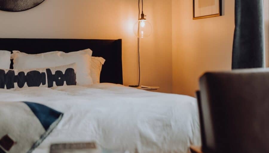 A neatly made bed with white linens and multiple pillows sits next to a small round side table and a hanging glass bulb lamp, casting a warm light in a modern, softly lit bedroom.