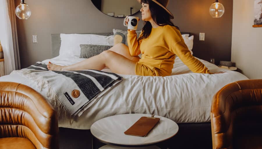 A woman in a yellow outfit sits on a bed with a striped blanket in a modern room. She holds a mug and wears a hat. Two pendant lights hang above, and there are two brown chairs and two round white tables in the foreground. A round mirror is on the wall.