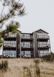 A four-story building with wooden siding and balconies is surrounded by dry grass. In the foreground, there are partially blurred green branches. The sky is overcast.