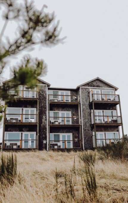 A four-story building with wooden siding and balconies is surrounded by dry grass. In the foreground, there are partially blurred green branches. The sky is overcast.