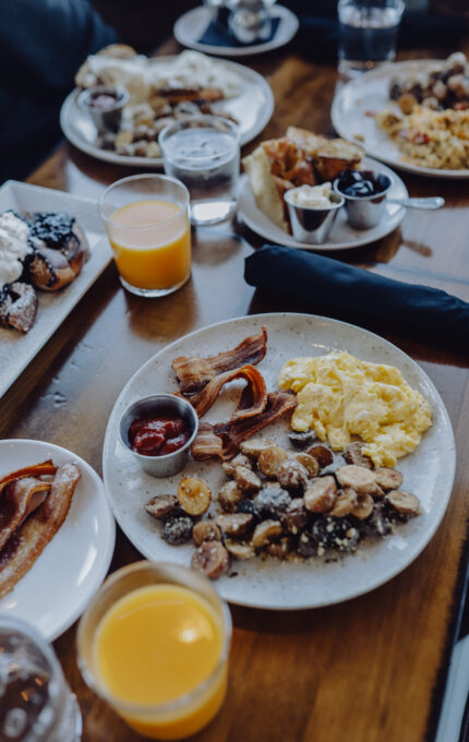 A table is set with plates of breakfast food, including scrambled eggs, bacon, and roasted potatoes. There are also glasses of orange juice and water. Additional dishes with various breakfast items are visible in the background.