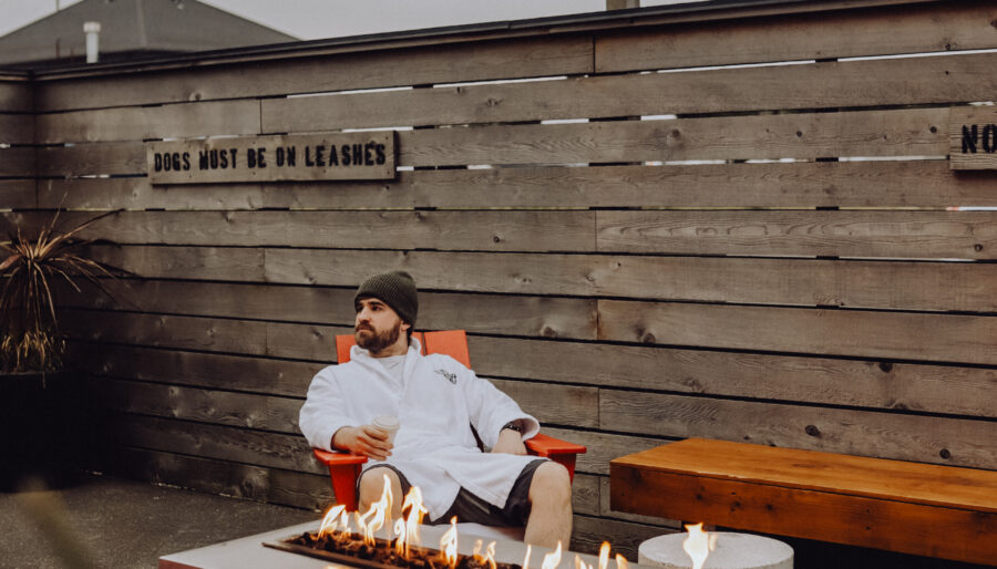 A person wearing a dressing gown and beanie sits in a red chair near a modern outdoor fire pit. Wooden benches and fairy lights surround the concrete patio. A wooden fence has a sign that reads "DOGS MUST BE ON LEADS.
