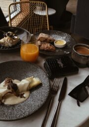 A table set with crepes topped with a creamy sauce, a dish of pastries with dipping sauce, a bowl with a dessert topped with blueberries and cream, a glass of orange juice, a mug, a wallet, sunglasses, and cutlery on a white marble surface.