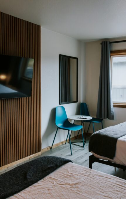 A hotel room with two beds, a wall-mounted TV, a small black table with two blue chairs by the window, a mirror on the wall, and a mini fridge in the corner. A building and trees are visible outside the window.