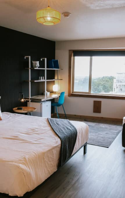 Modern hotel room with a double bed, pillows, and a brown cushion, a desk with a chair and lamp, two armchairs near a window, a TV mounted on the wall, and wood and dark accent decor.