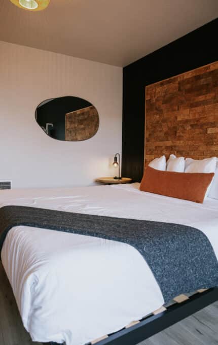 A neatly made bed with white linens, a gray throw blanket, and an orange accent pillow sits against a black and cork headboard. A wall lamp and oval mirror are mounted on the wall next to the bed.