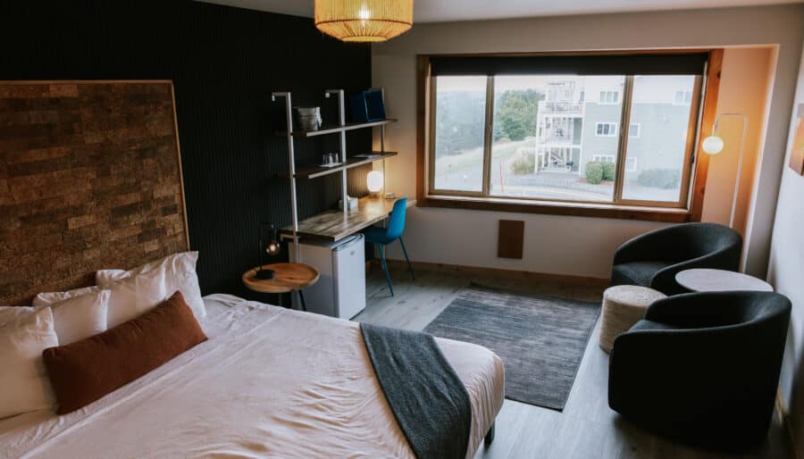 A modern hotel room with a bed, small desk, blue chair, mini fridge, two black lounge chairs, a round table, and a large window overlooking neighboring buildings and greenery.